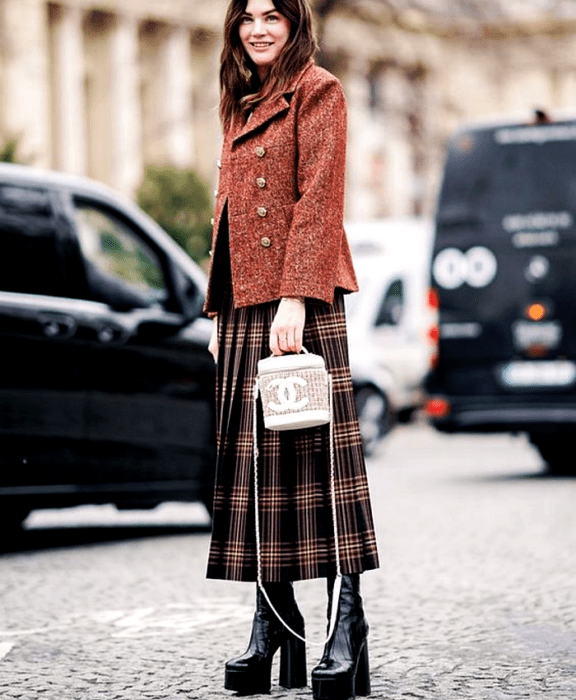 chica de cabello castaño usando un abrigo rojo con botones dorados, falda maxi de cuadros negros con rojo y café, botas de plataforma negras y bolso channel blanco bucketbag