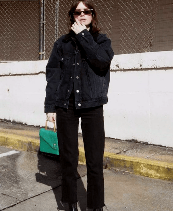 chica de cabello castaño usando lentes de sol, chaqueta de mezclilla negra, top negro, jeans negros rectos, botines negros, bolso de mano verde esmeralda