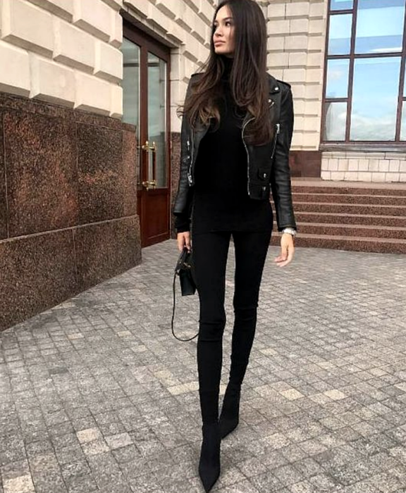 chica de cabello castaño usando un top negro de cuello alto, chaqueta de cuero negra, skinny jeans negros, botines de tacón negro, bolso de mano negro