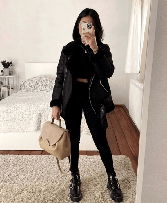 chica de cabello castaño usando lentes, crop top negro, chaqueta negra gruesa, skinny jeans negros, bolso de mano beige, botines de suela gruesa negros