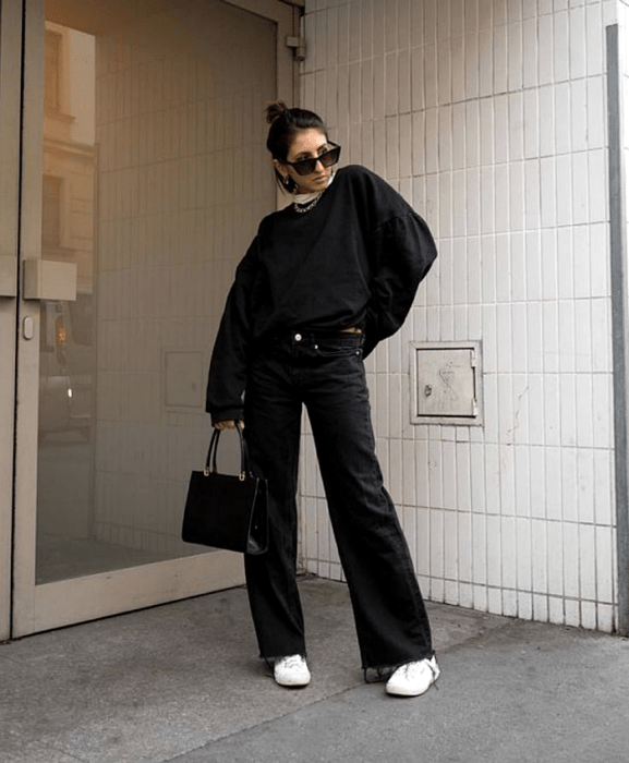 chica de cabello corto usando lentes de sol, sudadera negra oversized, jeans negros acampanados, tenis deportivos blancos, bolso negro de mano