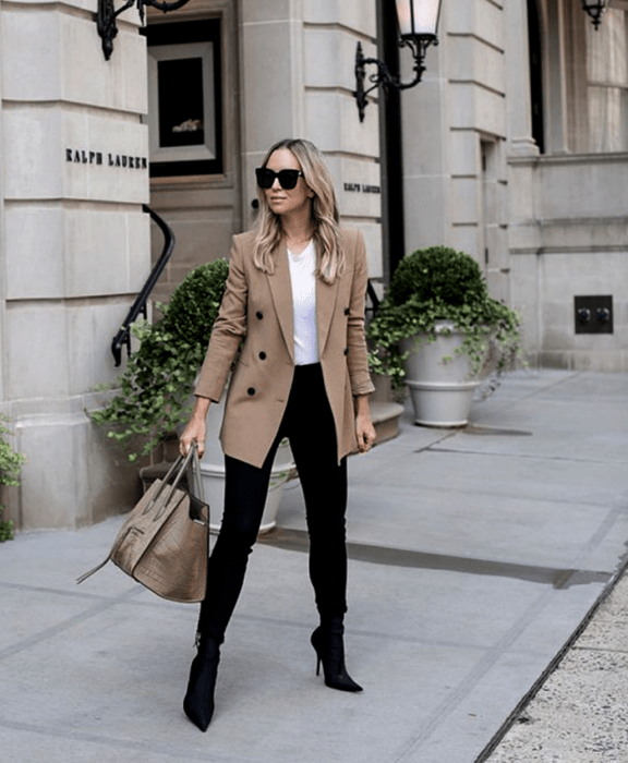 chica rubia usando lentes de sol, camiseta blanca lisa, blazer beige, skinny jeans negros, botines negros de tacón delgado y bolso de mano beige gris