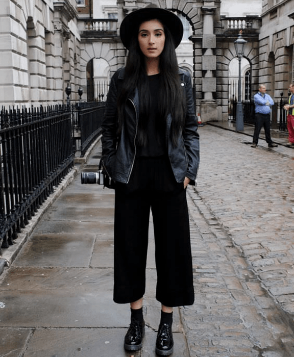 chica de cabello oscuro largo usando sombrero de ala ancha, camiseta negra, chamarra de cuero negra, jeans negros estilo culotte, botines de suela gruesa negros y cámara profesional negra