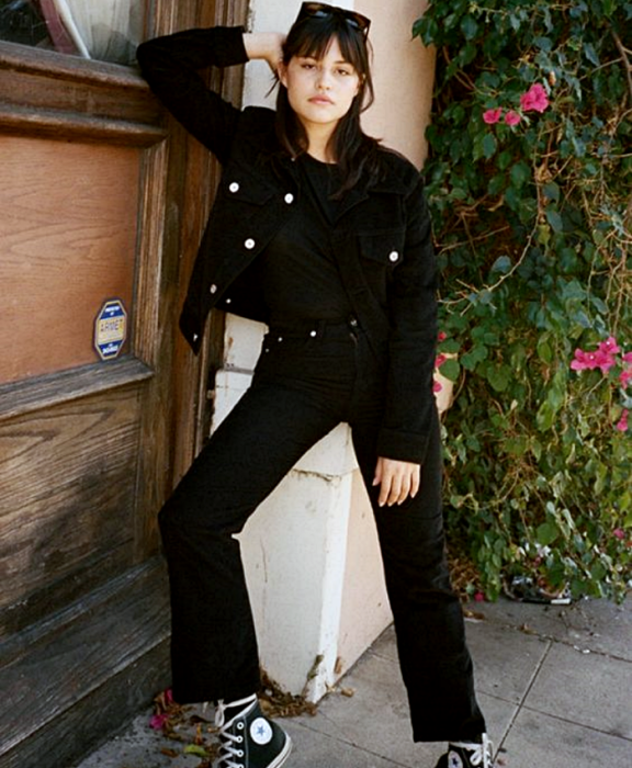chica de cabello castaño usando lentes de sol, top negro, chaqueta de mezclilla negra, jeans negros rectos y converse negros