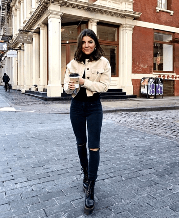 chica de cabello castaño usando una chaqueta de mezclilla beige, top negro de cuello alto, skinny jeans negros, botines de suela gruesa negros, café en mano