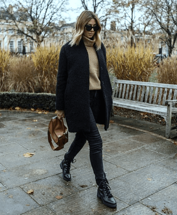 chica de cabello castaño claro usando lentes de sol, suéter beige de cuello alto, abrigo largo negro, skinny jeans negros, botines negros de suela gruesa y bolso de mano café