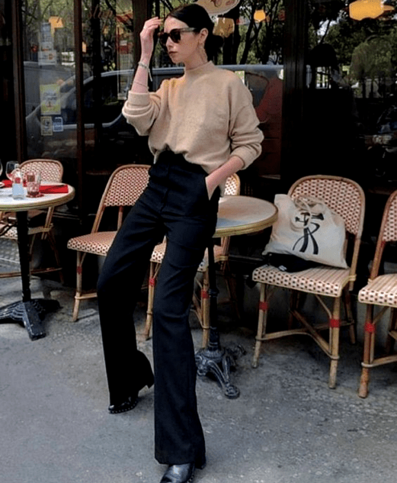 chica de cabello oscuro usando lentes de sol, suéter beige de cuello alto y manga larga, jeans acampanados negros, botines negros de tacón pequeño