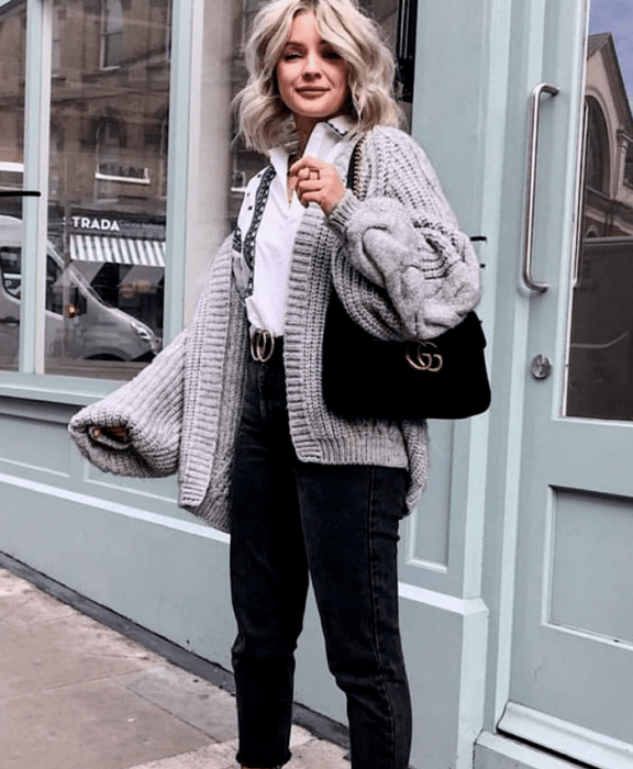 chica rubia platinada, usando una camisa blanca de vestir, cárdigan oversized gris, mom jeans negros, cinturón gucci y bolso de mano negro gucci