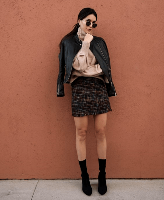 chica de cabello oscuro usando lentes de sol, suéter beige de manga larga, chaqueta de cuero, minifalda negra tejida con líneas blancas, botines estilo sock boots negras