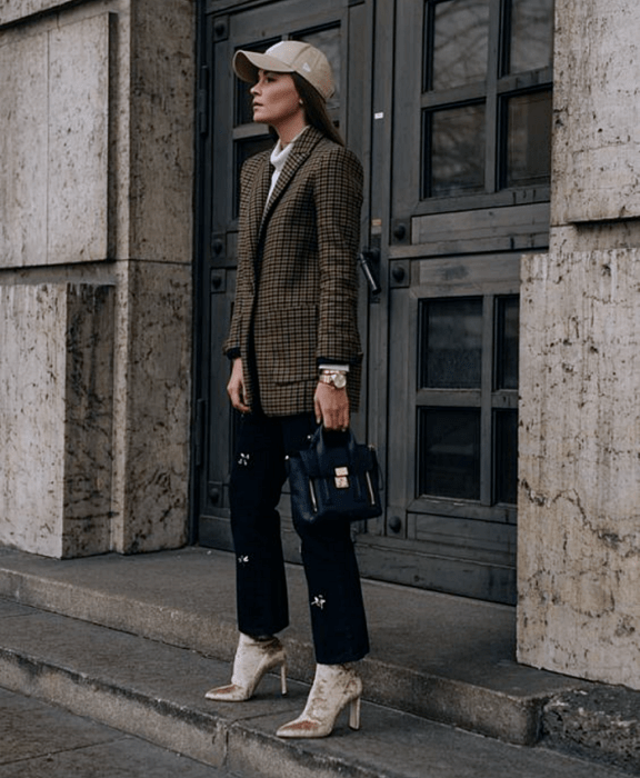 chica de cabello castaño usando una gorra beige, top blanco de cuello alto, blazer café de cuadros negros, pantalón negro de vestir con estrellas, botines beige de terciopelo estilo sock boot y bolso de mano negro