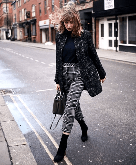 chica de cabello claro usando un top negro, abrigo negro con lunares blancos, pantalones grises de cuadros, bolso negro de mano y botines estilo sock boot negros