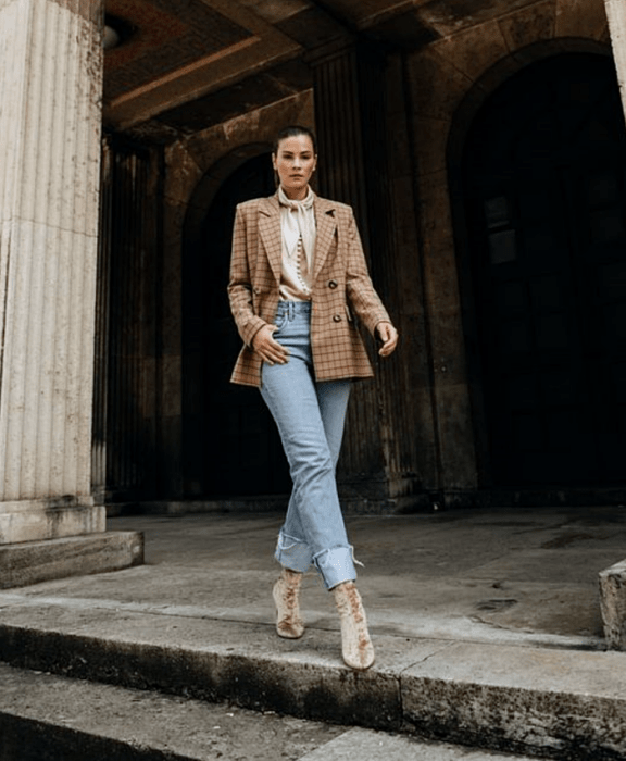 chica de cabello castaño usando una blusa beige de satén con moño en el cuello, blazer oversized beige de cuadros, jeans a la cintura estilo mom jeans, botines estilo sock boots beige de terciopelo