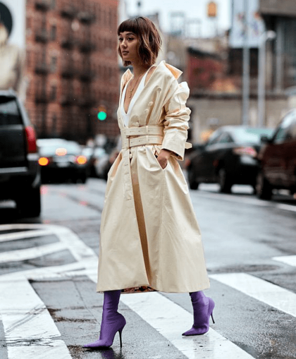 chica de cabello castaño corto usando un top de tirantes blanco, abrigo largo color beige, botas estilo sock boots moradas de satén