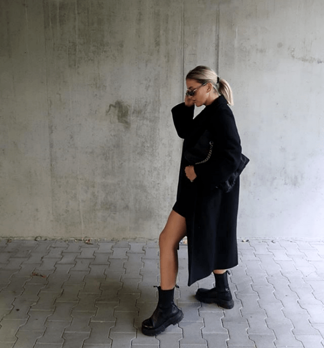 chica rubia con lentes de sol, usando un vestido negro corto, abrigo largo negro, botas negras de suela gruesa y bolso de mano negro