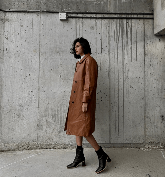 chica de cabello castaño usando un abrigo largo de piel café, botas de tacón negras con suela beige