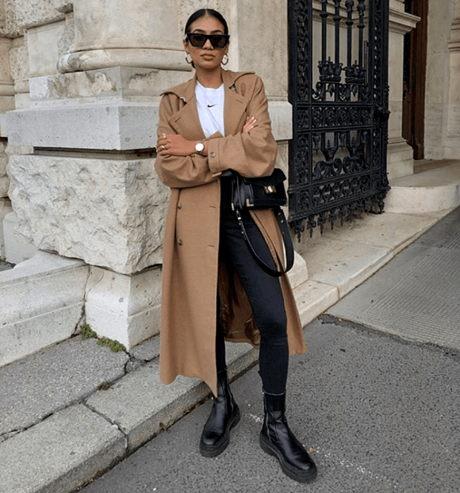 chica de cabello oscuro usando lentes de sol, camiseta blanca, abrigo largo café, skinny jeans negros, botas de cuero negras de suela gruesa y bolso de mano negro