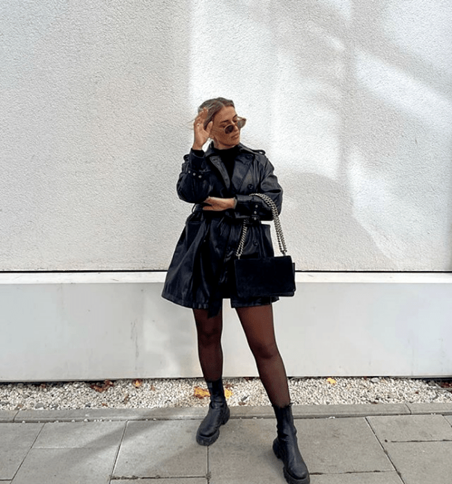 chica rubia usando lentes de sol, top negro de cuello alto, minifalda negra, abrigo negro de cuero, bolso de mano negro con correa de cadena, medias negras semitransparentes y botas de suela gruesa de cuero