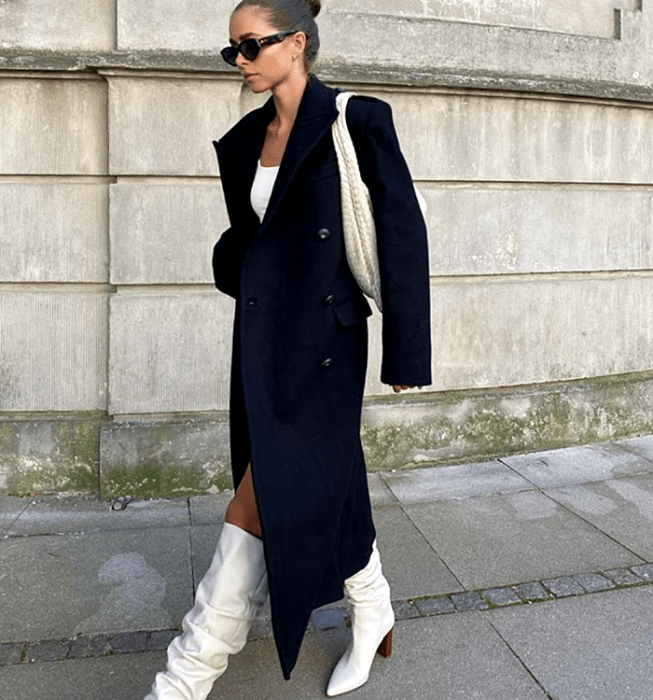 chica de cabello corto con lentes de sol, top blanco, abrigo largo negro, botas blancas de tacón café y bolso de mano blanco