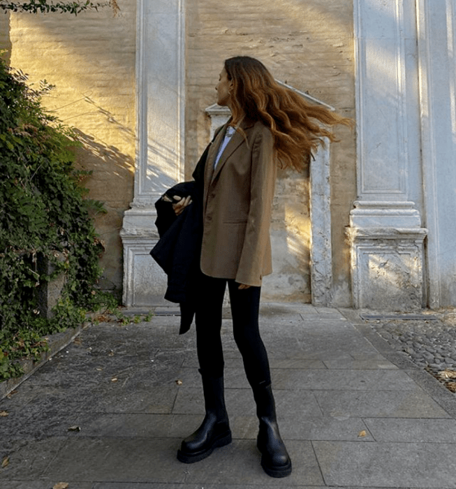 chica de cabello claro usando una camiseta blanca, abrigo beige oscuro, skinny jeans negros, abrigo negro y botas de suela gruesa negras