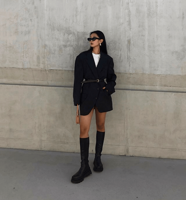 chica de cabello oscuro con lentes de sol, camiseta blanca, abrigo oversized negro con cinturón, botas largas de plataforma gruesa