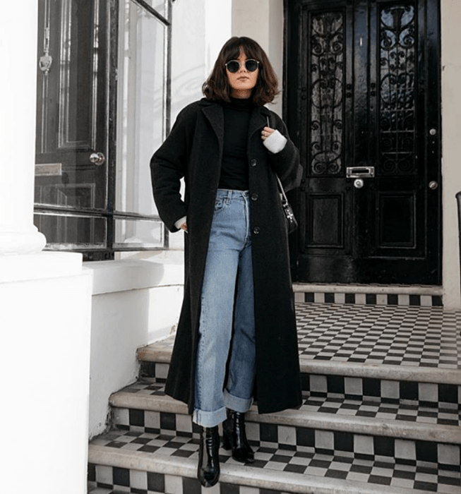 chica de cabello castaño oscuro usando lentes de sol, top negro de manga larga, abrigo largo negro, jeans a la cintura rectos, botas negras de tacón y bolso negro de mano