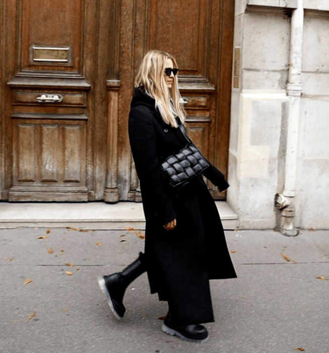 chica rubia con lentes de sol, usando un top negro, leggings negros, abrigo largo negro, bolso de mano negro de piel y botas de suela gruesa negras