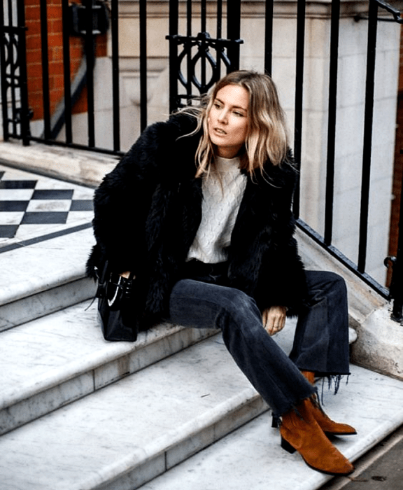 chica rubia usando un suéter blanco, abrigo afelpado negro, jeans oscuros, bolso de mano negro y botines cafés de terciopelo ante