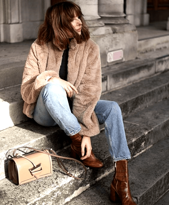 chica de cabello castaño usando un abrigo afelpado café, top negro, jeans azules, botines de piel cafés de tacón, bolso de mano beige