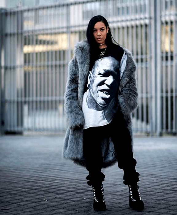 chica morena de cabello oscuro usando una camiseta oversized negra con estampado, abrigo afelpado gris, pantalones negros holgados, tenis de plataforma negros con agujetas