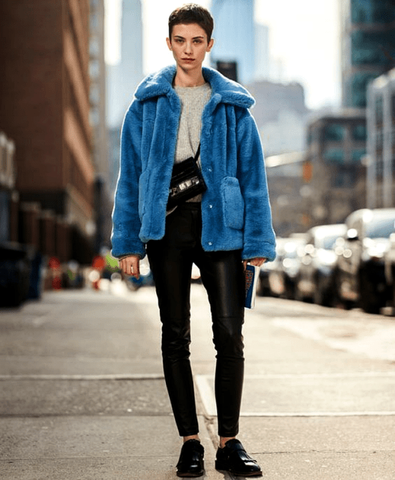 chica de cabello corto usando un suéter gris, abrigo afelpado azul, bolso negro crossover, pantalones de cuero negro, zapatos loafers negros