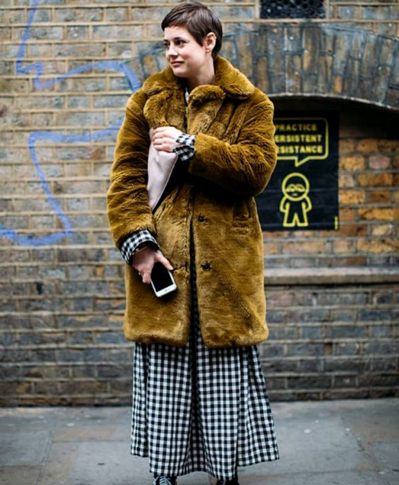 chica de cabello corto usando abrigo afelpado color mostaza, vestido maxi de cuadros blancos, vans negros y bolso beige de mano