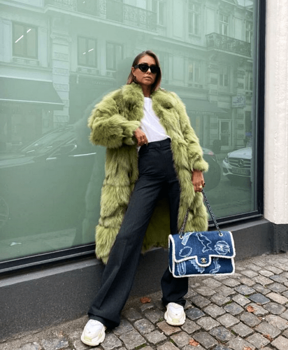 chica rubia usando lentes de sol, camiseta blanca, pantalones de vestir a la cintura color gris oscuro, tenis deportivos blancos, bolso de mezclilla grande y abrigo afelpado verde