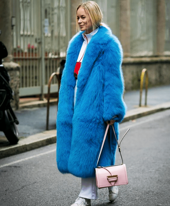 chica rubia usando una sudadera blanca con vistas rojas, abrigo afelpado azul brillante, pantalones blancos, tenis deportivos blancos y bolso de mano rosa pastel