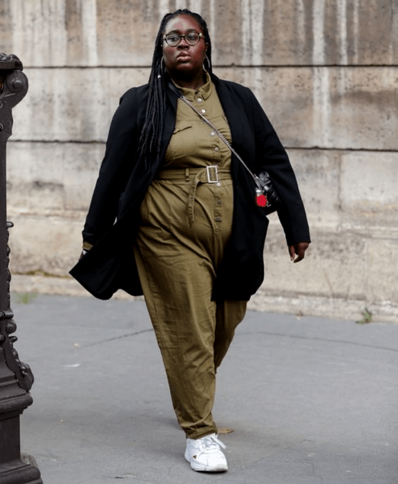 chica morena curvy usando lentes, jumpsuit verde militar con ajuste a la cintura, abrigo negro largo, bolso crossbody bag negro de piel y tenis deportivos blancos