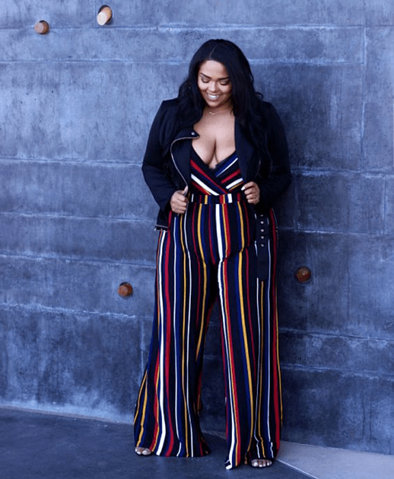 chica morena curvy de cabello oscuro usando un jumpsuit de rayas de colores azul, blanco, rojo y amarillo, con escote pronunciado en V y chaqueta negra