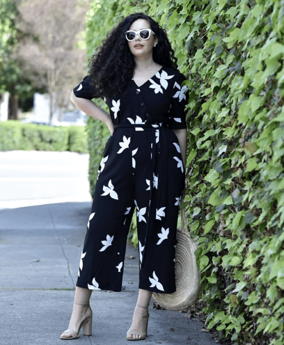 chica curvy de cabello chino usando lentes blancos de sol, jumpsuit negro con flores blancas, con escote en V y ajuste en la cintura, sandalias nude de tacón y bolso de rafia redondo de mano