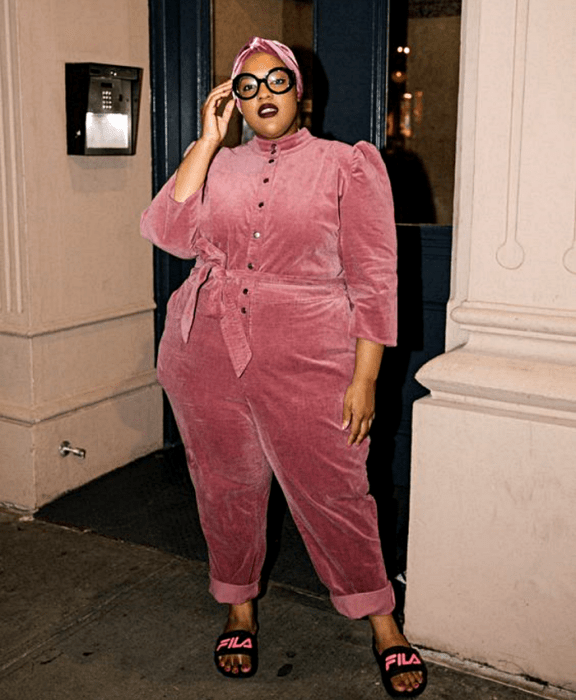 chica morena curvy usando lentes, banda rosa en el cabello, jumpsuit rosa de terciopelo con ajuste en la cintura, manga larga y pantalón largo, con sandalias negras fila