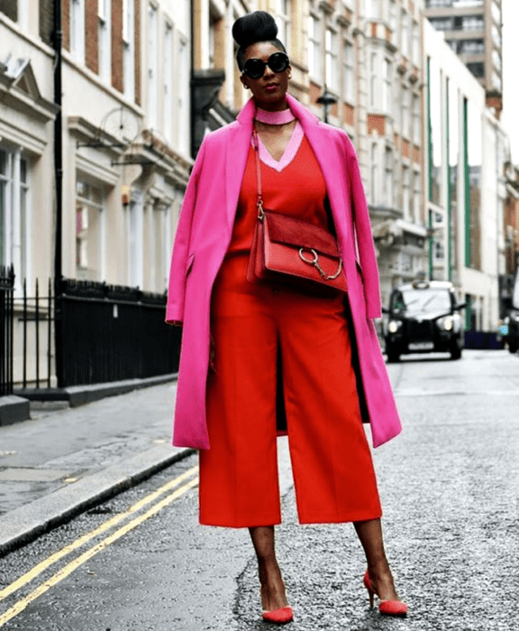 chica morena curvy usando lentes de sol, jumpsuit rojo con cuello en v, abrigo rosa largo, bolso crossbody bag rojo de piel con zapatos de tacón rojos