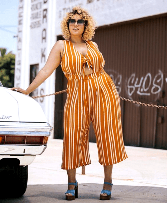 chica rubia curvy usando lentes de sol, jumpsuit naranja sin mangas con escote en v, líneas blancas, ajustado a la cintura con pantalón tipo culotte y zapatos de tacón azul