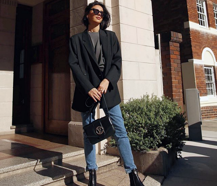 chica de cabello corto usando lentes de sol, top gris, blazer oversized negro, skinny jeans, botines negros y bolso negro de piel