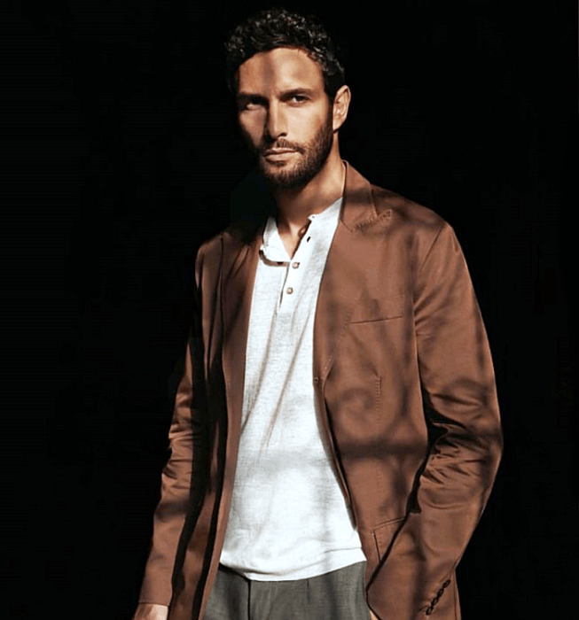 Noah Mills; chico de cabello castaño usando una camiseta blanca con botones en el cuello, blazer café largo y pantalón de vestir café oscuro