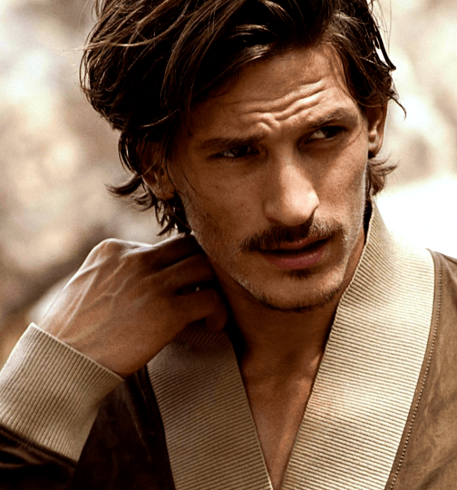 Jarrod Scott; chico de cabello castaño con bigote usando un cárdigan café con cuello beige 