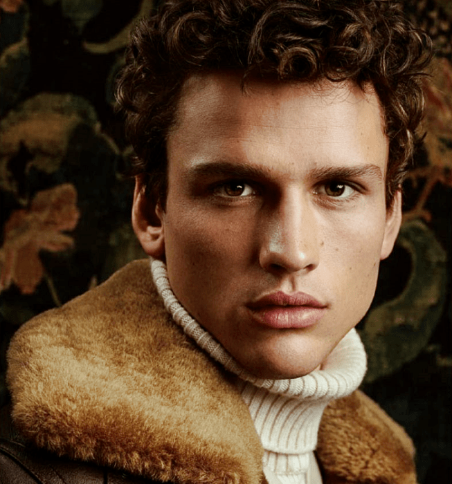 Simon Nessman; chico de cabello chino café usando un suéter beige de cuello alto y chamarra café con cuello afelpado