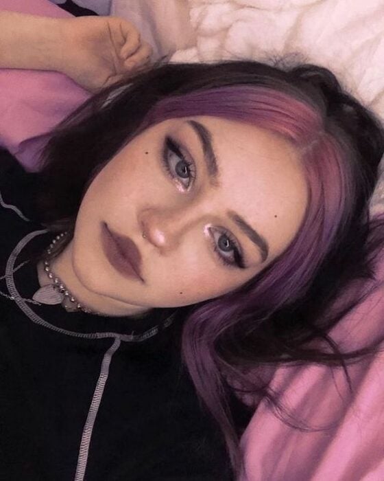 Chica acostada en su cama con cabello con mechones morados, labial morado y delineado
