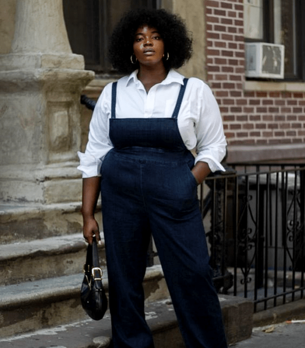 chica curvy morena de cabello chino usando una camisa de vestir blanca, overall overol de mezclilla, bolso negro de mano