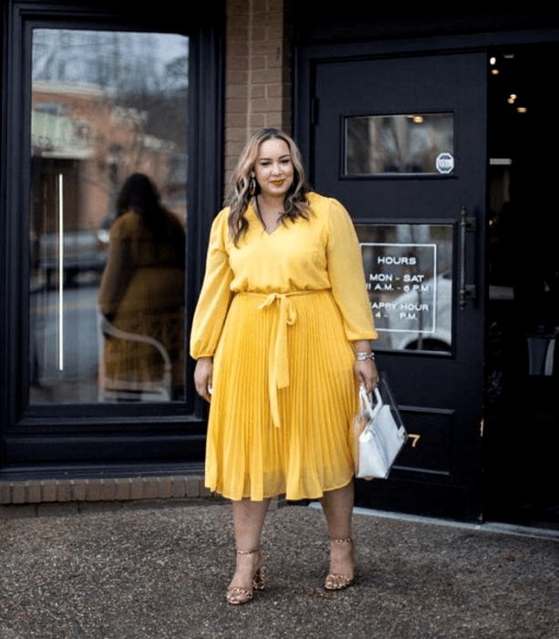 chica curvy de cabello castaño claro usando un vestido amarillo de manga larga, con ajuste en la cintura y falda larga, sandalias de tacón de animal print y bolso de mano blanco