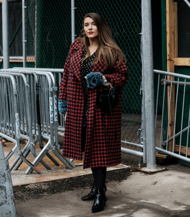 chica curvy de cabello castaño usando una blusa negra de lunares blancos, leggings negros, botines negros de piel con tacón y bolso negro de mano