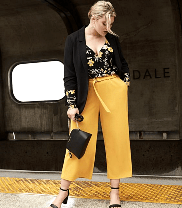 chica rubia usando un top negro de flores, pantalón de vestir amarillo, tacones negros y bolso negro de mano