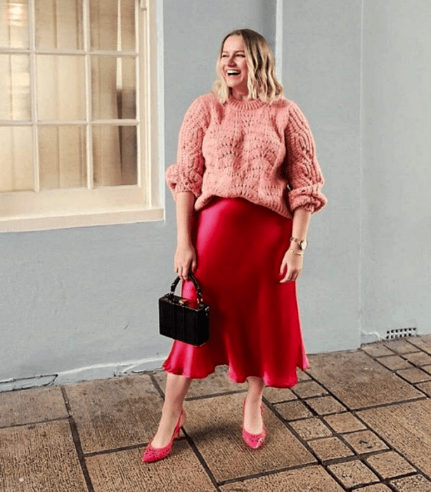 chica rubia curvy usando un suéter oversized rosa, falda de satén rosa fucsia, zapatos de tacón rosas y bolso de mano negro