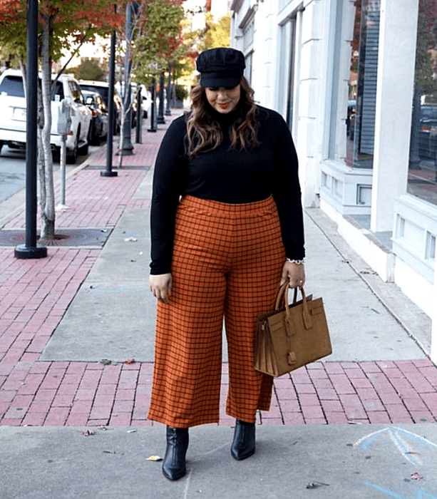 chica curvy de cabello castaño claro usando un gorro negro, suéter negro de cuello alto, pantalones naranjas con lineas negras, botines de piel negras de tacón y bolso café de mano